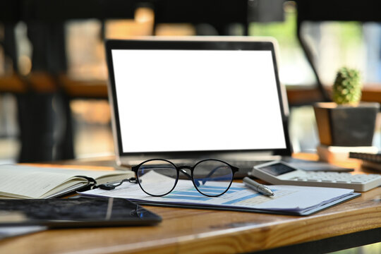 Photo of a comfortable working desk surrounded by a white blank screen computer laptop, eyeglasses, calculator, pen, notebook, digital tablet and document.