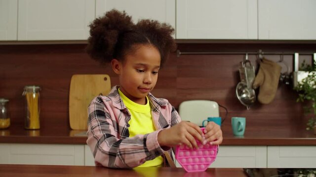 Lovely focused elementary age African American girl with trendy hairstyle playing with sensory pop it fidget toy, relaxing and soothing anxiety and stress while child relaxing in domestic kitchen.