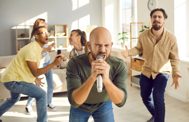 Obraz premium Karaoke at home. Emotional young man holding a microphone and enthusiastically singing while having fun with friends at home. Man sings against background of friends who also sing and have fun in room
