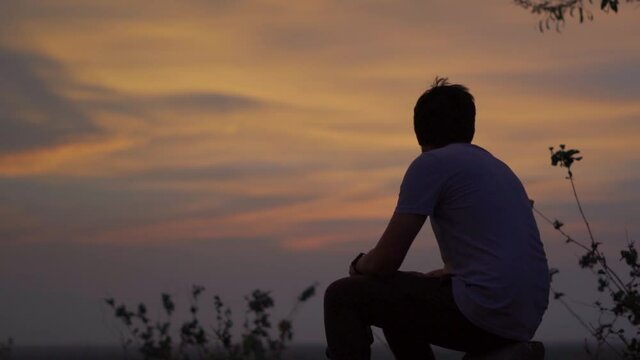 Silhouette shot of an Indian man sitting in front of the orange clouds during the sunset. Man lost in deep thoughts in front of the sunset. Loneliness background