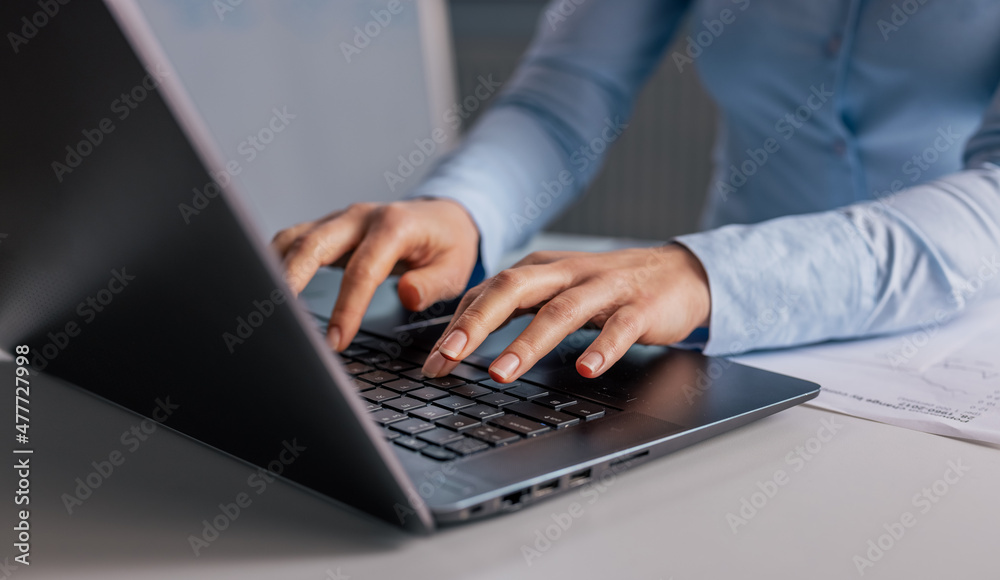 Wall mural Close up of mature woman in blue shirt typing on keyboard of modern laptop. Business lady sitting at office desk and using portable computer for work.