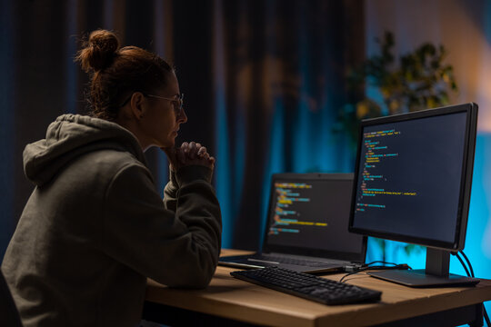 Thoughtful woman in casual wear and eyeglasses sitting at living room during evening time and looking on two modern computers. Competent programist typing data code at home.