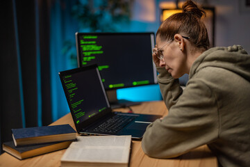 Side view of focused caucasian woman in eyewear sitting at desk with laptop and pc while examining html code on screens. Late work at home of female programmer.