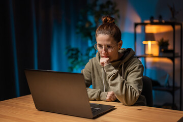 Mature woman in eyewear sitting at desk and looking on laptop screen with thoughtful facial expression. Female freelancer working at night while staying at home.