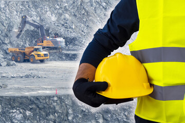 A worker holds a construction helmet in his hand against the background of a mining quarry, in which an excavator and a heavy dump truck are working