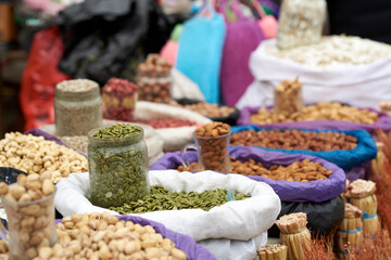 A glass jar of green raw pumpkin seeds is in a white sack. There are sacks of nuts around - pistachios, cashews, almonds and others. Blurred background, artistic bokeh effect.