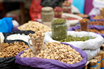 A glass cup with inshell pistachios is in a lilac sack on the market. Around are bags of walnuts, green pumpkin seeds, cashews, almonds and other nuts. Blurred background, artistic bokeh effect.