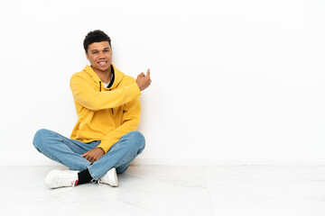 Young African American man sitting on the floor isolated on white background pointing back