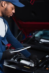 Fototapeta na wymiar young african american repairman holding digital tablet while making inspection of blurred car.