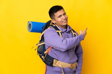 Young mountaineer man with a big backpack isolated on yellow background pointing back