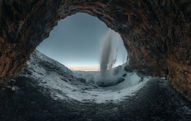 View of Seljalandsfoss waterfall at dawn in winter from behind in Iceland with snow and frozen ice in the foreground.