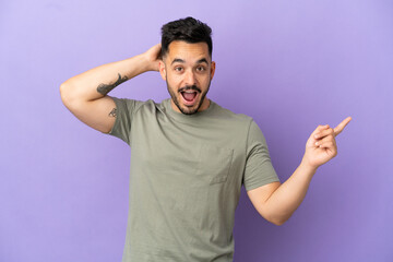 Young caucasian man isolated on purple background surprised and pointing finger to the side
