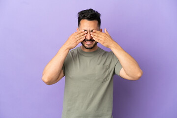 Young caucasian man isolated on purple background covering eyes by hands and smiling