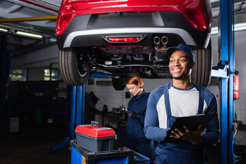 Young african american mechanic holding clipboard near colleague and car in service.