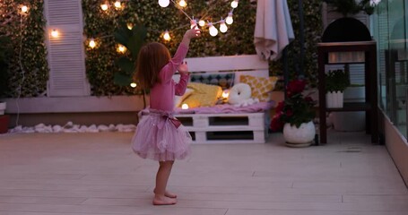 cheerful toddler baby girl having fun on rooftop patio at summer evening, playing with party light bulb strings