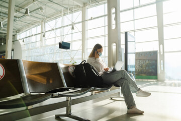 Female traveler in mask sits on chair in airport lobby with a social distance sign, talking on the phone and working on her laptop