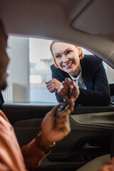 cheerful car dealer giving key to blurred african american buyer sitting in car.