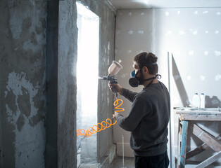 a male plasterer in a construction respirator paints a wall from a spray gun before plastering and...