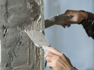 a plasterer man with a beard stands at a rough plastered wall with spatulas. 
