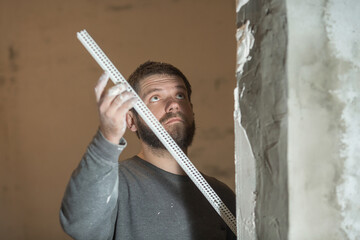 a man with a beard plasterer with the help of holds.a perforated corner and looks intently at the...