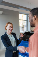 happy car dealer holding key and shaking hands with blurred african american customer.