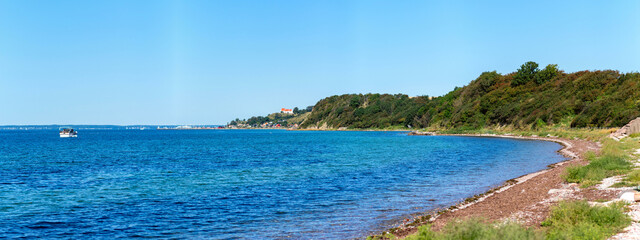 Obraz premium Panorama of the coast of Ven, an island of the coast of Landskrona, Sweden. Calm summer sea, blue water and sky. Sunlit coastline on the other side. Beach with trees and gravel