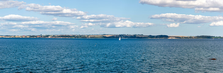 Fototapeta premium Panorama of the coast of Ven, an island of the coast of Landskrona, Sweden. Calm summer sea, blue water and sky. Sunlit coastline on the other side. Beach with trees and gravel