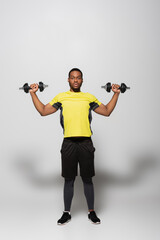 Fototapeta na wymiar full length of african american sportsman working out with heavy dumbbells on grey.