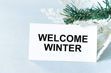 Welcome Winter text on a card on a light background next to spruce branches, festive background