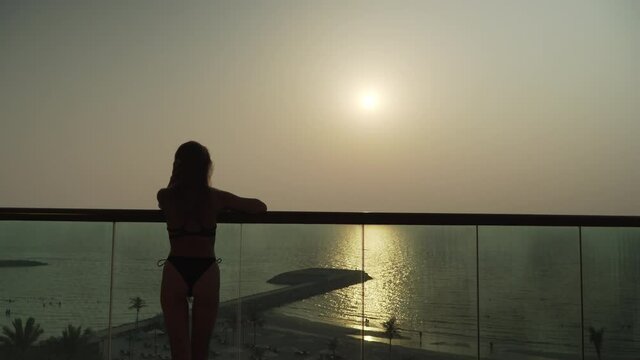 A slender beautiful Caucasian girl in a swimsuit raises her hands up. A European woman stands on the balcony against the backdrop of the sunset and the sea. Lady in bikini on vacation. evening 