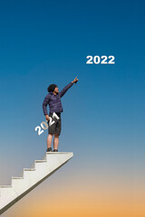 Young man holding a letter representing the year 2021 standing on the top of the white ladder that extends up into the sky. He pointed his index finger to the sky where the year 2022 characters appear