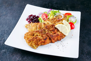 Traditionelles frittiertes Schnitzel mit grünen Salat, Tomaten und Preiselbeeren serviert als close-up auf einem Design Teller mit Textfreiraum
