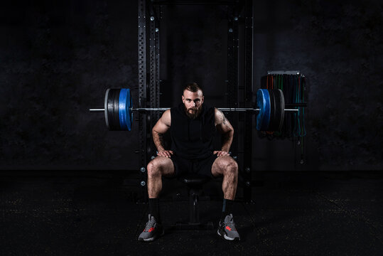 Young active strong sweaty muscular fit man with big muscles preparing for hardcore benchpress workout cross training with heavy barbell weights in the gym. Male concentrated sitting on the bench.