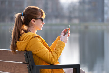 Young pretty woman sitting on a park bench browsing her smartphone outdoors on warm autumn day
