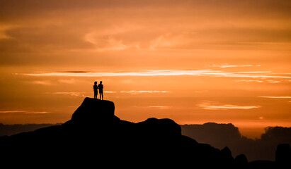 Dramatic golden sunset of two men admiring the view on vacation, standing on a large rock on a beach in Jacobsbaai, South Africa.