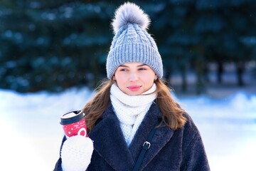 Portrait of beautiful attractive girl, happy positive joyful pretty woman in warm clothes, knitted hat and scarf at winter frosty cold snowy day is holding, drinking cup of hot tea or coffee, smiling