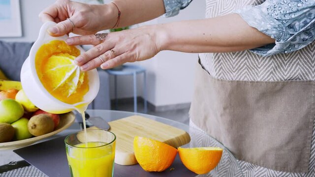 Unrecognizable lady in grey apron pours fresh orange juice into cup from squeezer at glass table in light contemporary kitchen. Woman prepare healthy breakfast in morning