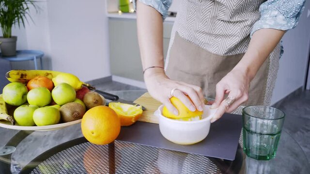Unrecognizable lady in grey apron pours fresh orange juice into cup from squeezer at glass table in light contemporary kitchen. Woman prepare healthy breakfast in morning