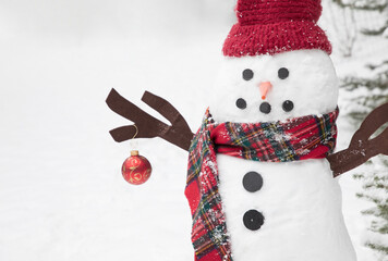An original wintery Christmas themed image of a snowman dressed in red tartan plaid holding a Christmas ornament in the snow