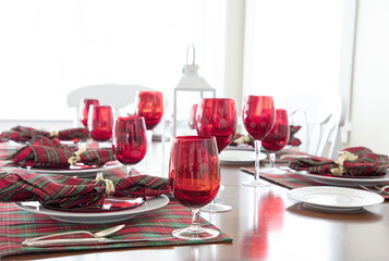 An original lifestyle photograph of a festive holiday dinner table set with red glassware and tartan plaid Christmas napkins against a white background.