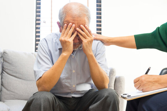 Depressed Asian senior man sharing problems during therapy session with female psychiatrist writing on clipboard.