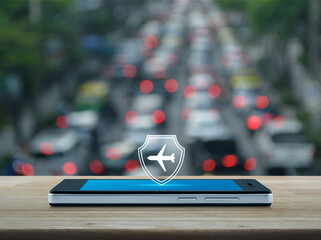 Airplane with shield flat icon on modern smart mobile phone screen on wooden table over blur of rush hour road in city, Business travel insurance and safety online concept