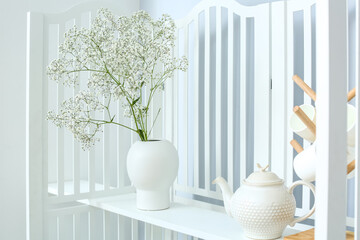 Vase with gypsophila flowers, teapot and cups on folding screen in light room