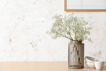Vase with gypsophila flowers, cup and bowls on table near light wall
