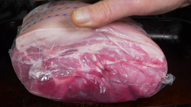 Closeup of a man&rsquo;s hand placing a fresh, raw, pork joint with its blood juices, on a baking tray, ready to be unsealed from its plastic packaging.