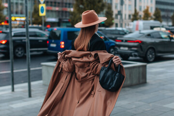 Young blonde woman in beige hat and long autumn brown leather coat walking in the city. Street...