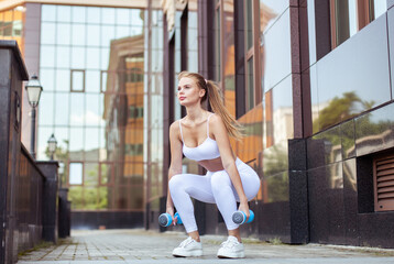 Young red-haired fit woman with freckles squats with dumbbells in the city. Healthy lifestyle. Outdoor workout