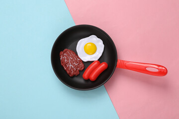 Toy frying pan with scrambled eggs, meat, sausage on pink blue background