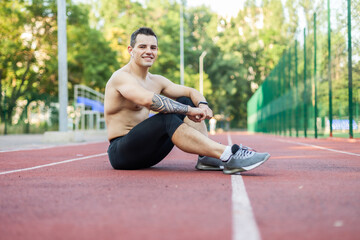 Fototapeta na wymiar Smiling young athlete man runner with naked torso resting while sitting on stadium