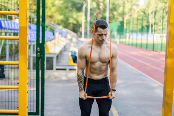 Athletic male athlete with a naked torso trains with a fitness elastic band on the workout site. Outdoor workout